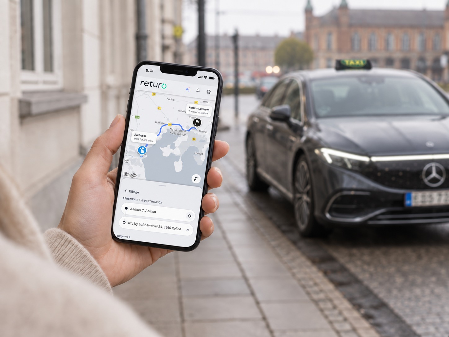 En hånd holder en telefon, hvor Returo-appen viser ruten til Aarhus Lufthavn, foran en taxa